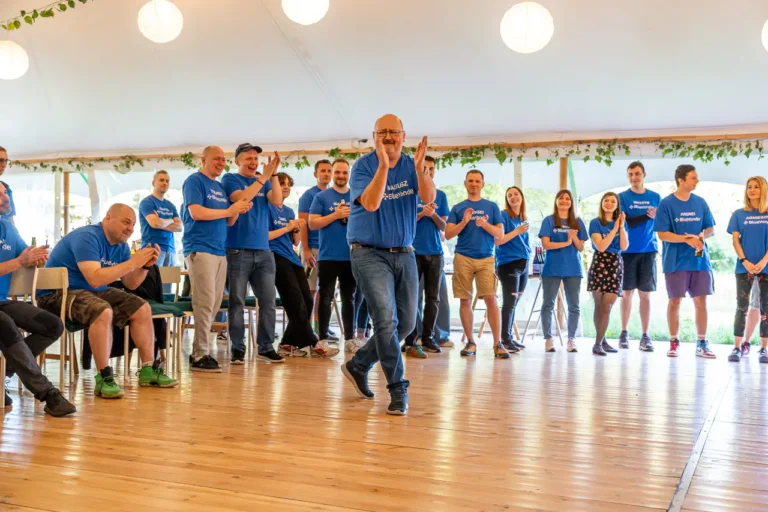 Grupa osób ubranych w identyczne, niebieskie koszulki stoi w półkolu na drewnianej podłodze, klaszcze i uśmiecha się, gdy jedna osoba idzie do przodu, do środka, pod udekorowanym namiotem — to idealna sceneria dla fotografii firmowej.