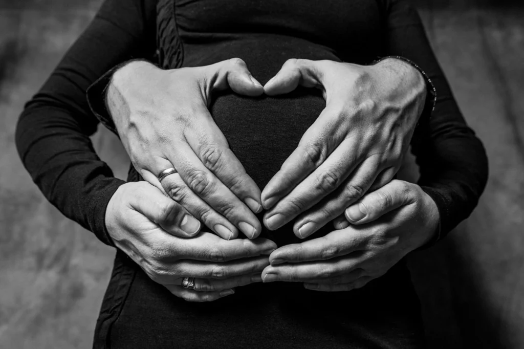 Czarno-białe zdjęcie autorstwa Fotograf Rzeszów przedstawia dwie pary dłoni, z których jedna nosi obrączkę ślubną, delikatnie spoczywające na brzuchu kobiety w ciąży. Górne dłonie tworzą kształt serca, symbolizującego miłość i oczekiwanie.