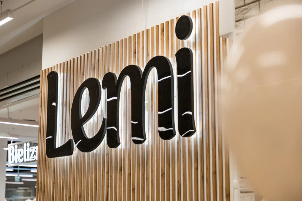Znak ścienny z napisem Lemi z dużymi, czarnymi, zaokrąglonymi literami jest zamontowany na pionowych drewnianych listwach w dobrze oświetlonej przestrzeni wewnętrznej - idealnej do profesjonalnych zdjęć i oddania atmosfery otwartego sklepu Rzeszów.