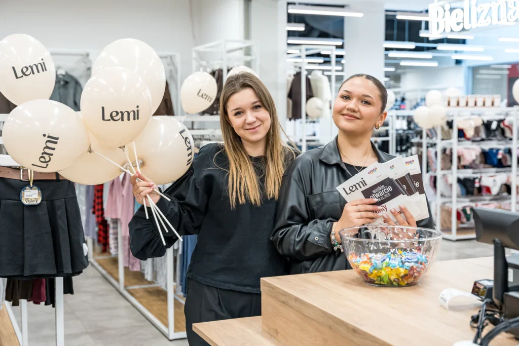 Dwie młode kobiety stoją za ladą w otwartym sklepie w Rzeszowie. Jedna trzyma beżowe balony "Lemi", podczas gdy druga rozdaje broszury. Na ladzie stoi szklana miska z kolorowymi cukierkami, uchwycona na tych profesjonalnych zdjęciach.