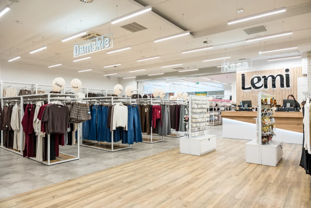 Wnętrze sklepu odzieżowego z wieszakami z odzieżą damską, dobrze oświetlonym, nowoczesnym wystrojem, drewnianą podłogą i kasą z napisem Lemi. Widoczne są szyldy Damskie i Kasa - profesjonalne zdjęcia z otwarcia sklepu Rzeszów.
