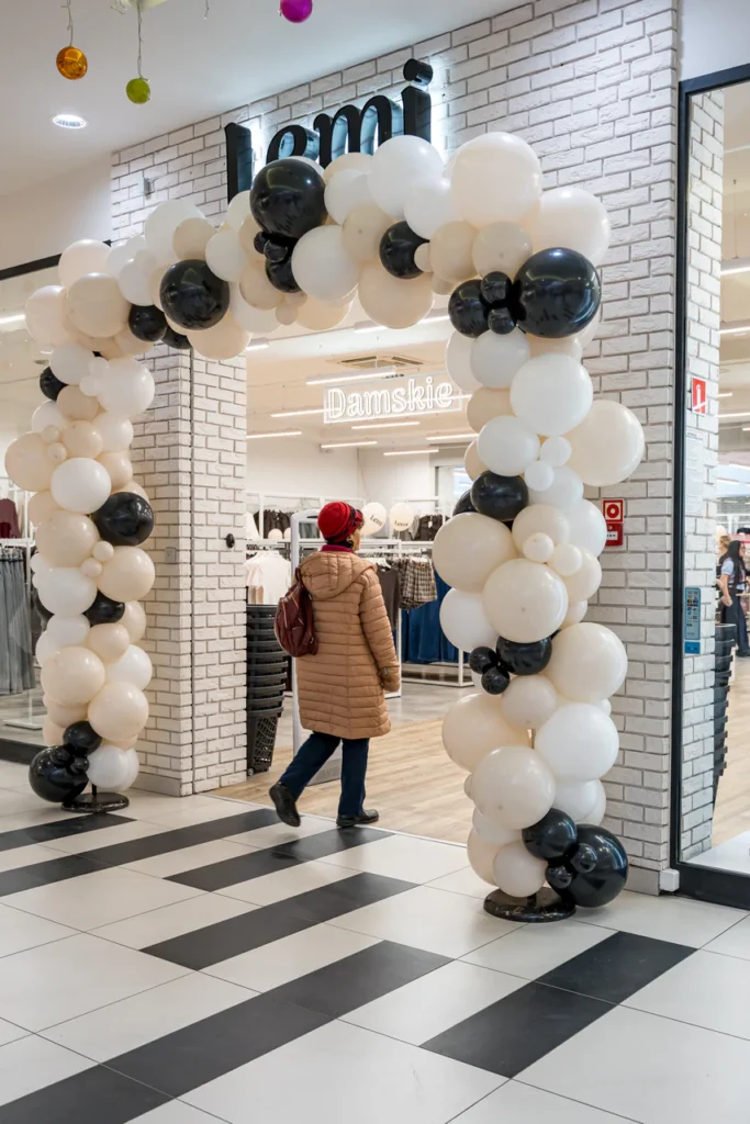 Osoba w brązowym płaszczu i czerwonym kapeluszu przechodzi przez duży łuk czarno-białych balonów na otwarciu sklepu Rzeszów, uchwycona na profesjonalnym zdjęciu przy wejściu do sklepu odzieżowego w galerii handlowej.