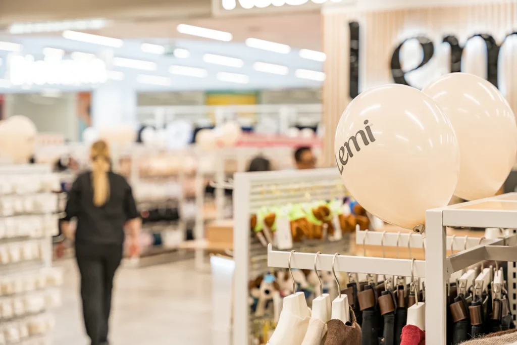 Wnętrze sklepu detalicznego z wieszakami odzieżowymi i białym balonem z napisem "emi" w centrum uwagi, uchwycone podczas otwarcia sklepu Rzeszów. Osoba o blond włosach odchodzi w jasno oświetlonej, zorganizowanej przestrzeni - idealnej do profesjonalnych zdjęć.