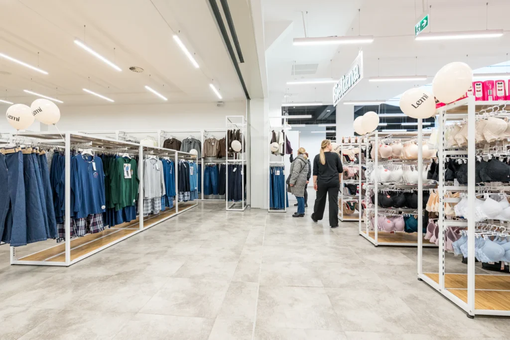 Wnętrze sklepu odzieżowego z wieszakami z dżinsami, koszulami, kurtkami i biustonoszami. W tle przeglądają się trzy osoby. Podłoga wyłożona kafelkami i czysta, dobrze oświetlona przestrzeń tworzą przyjazną atmosferę - idealną do profesjonalnych zdjęć podczas otwarcia sklepu w Rzeszowie.