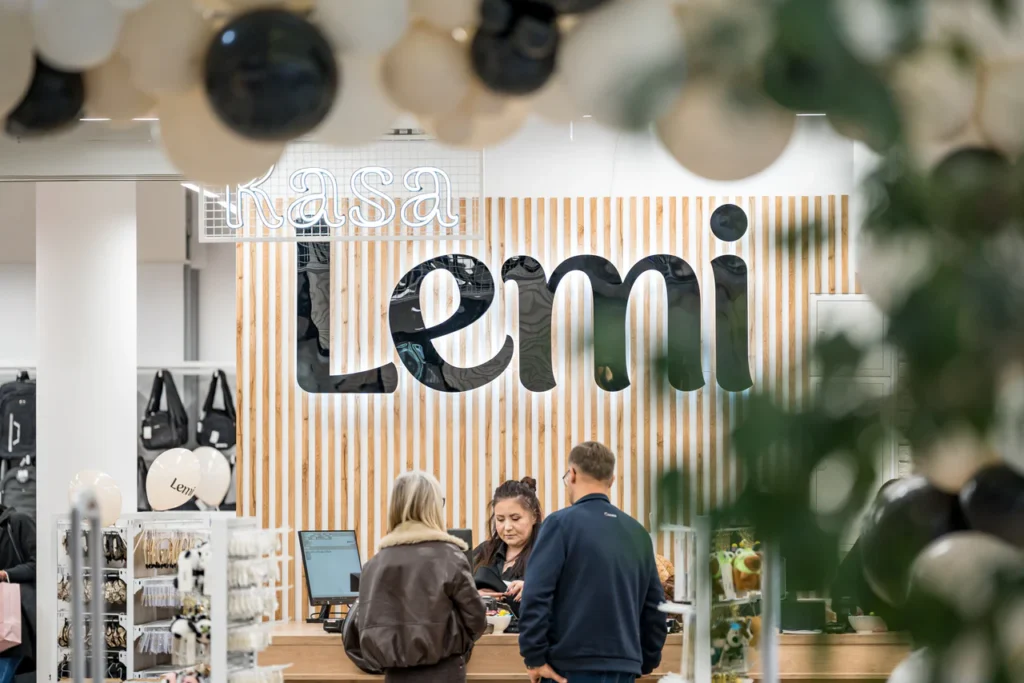 Trzy osoby stoją przy ladzie sklepowej w pobliżu kasjera, z dużym szyldem Lemi i drewnianą ścianą w paski w tle. Ozdobne balony widoczne są na pierwszym planie podczas otwarcia sklepu Rzeszów, uchwycone na profesjonalnym zdjęciu fotorelacja.
