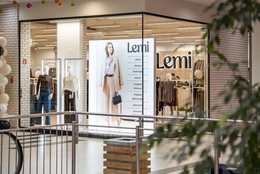 Witryna sklepu odzieżowego Lemi w centrum handlowym podczas otwarcia sklepu w Rzeszowie, z dużym plakatem przedstawiającym kobietę w beżowym garniturze i różnymi ubraniami wyeksponowanymi wewnątrz. Na pierwszym planie widoczna jest część balustrady i zieleń.