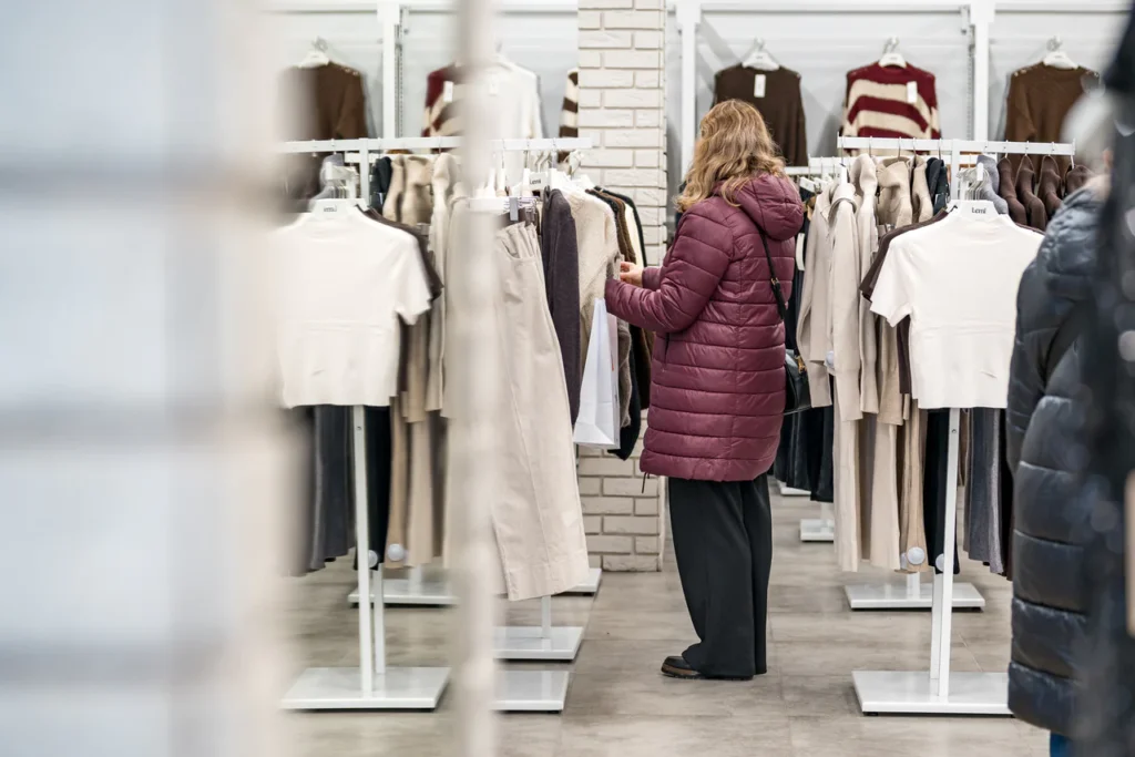 Osoba ubrana w bordową kurtkę przegląda wieszaki z odzieżą w sklepie wypełnionym neutralnymi i ziemistymi ubraniami, uchwyconymi na profesjonalnych zdjęciach, które podkreślają zachęcającą atmosferę tego wydarzenia otwarcie sklepu Rzeszów.