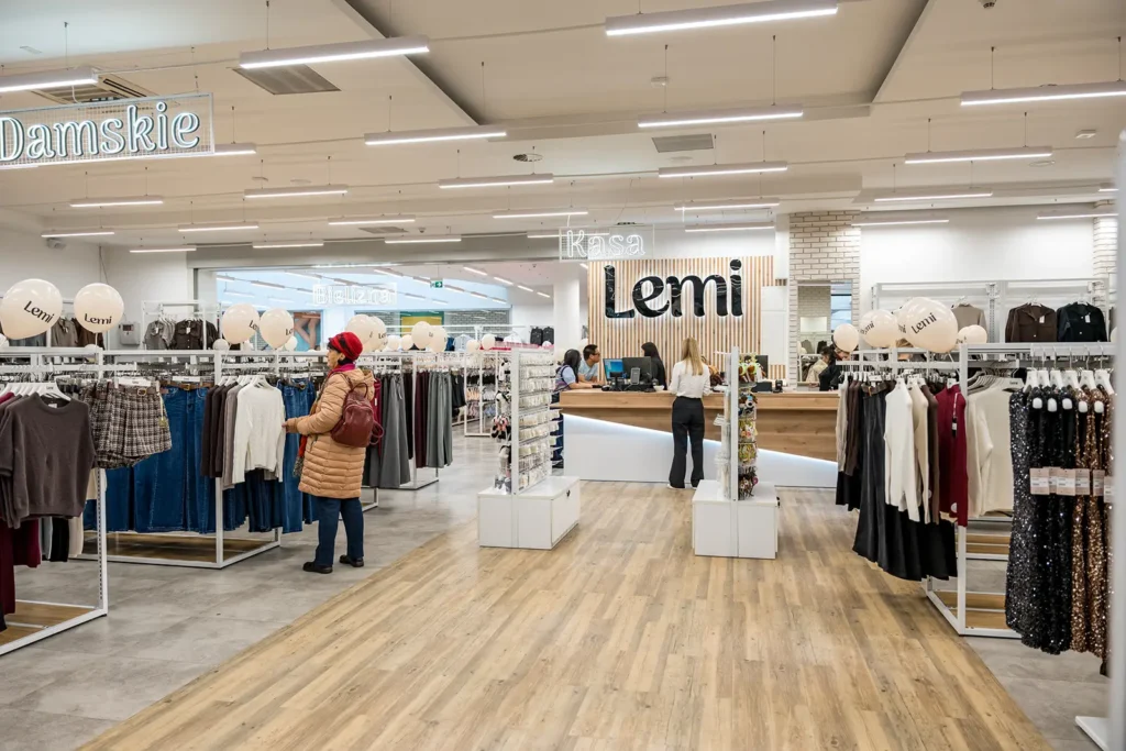 Wnętrze sklepu odzieżowego z wieszakami z damską odzieżą, drewnianą podłogą i kupującymi. Profesjonalne zdjęcia przedstawiają otwarcie sklepu w Rzeszowie.